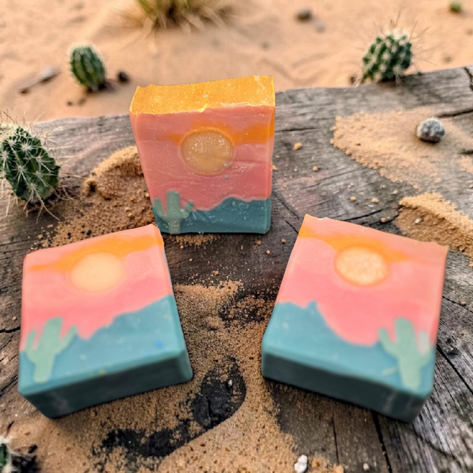 Three rectangular handmade soap bars with a desert sunset design, featuring a yellow sun, pink sky, and teal mountains with cacti, displayed on a weathered wooden surface outdoors with sand and cacti in the background.