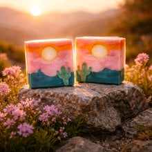 Two rectangular layered soap bars with a desert sunset design, featuring pink and orange sky, a yellow sun, teal mountains, and green cacti, displayed on a rock outdoors at sunset.
